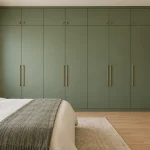 Bedroom with a full-height matte sage-green hinged wardrobe, slim brass handles, matching overhead loft storage, light oak floor, cream bed partly visible. Entire wardrobe visible.