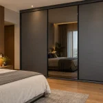 Contemporary bedroom with a charcoal-grey sliding wardrobe with one smoked mirror panel, slim black profiles, warm oak flooring and soft bedside lighting. Entire wardrobe visible.